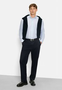 Herrenoutfit mit einem hellblauen Hemd mit Knöpfen, einem marineblauen Pullover, der über den Schultern liegt, dunklen Hosen und schwarzen Schuhen.