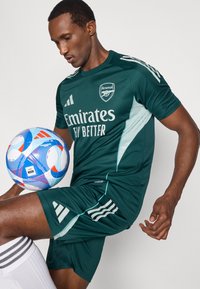 Grønt Arsenal fodboldudstyr med korte ærmer og lodrette striber, sammen med en flerfarvet fodbold, placeret foran en ensfarvet baggrund.