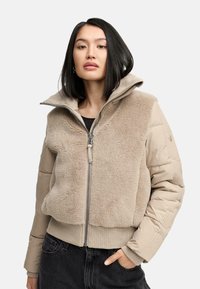 Beige fleece en gewatteerd jack met een hoge kraag, ritssluiting en geribbelde manchetten. Het heeft een zachte, gestructureerde voorkant en gladde mouwen.