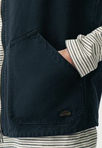 Marineblauwe canvas vest met een rits aan de voorkant, met een schuine zak en een klein merkembleem. Het binnenste shirt heeft horizontale marineblauwe en witte strepen.