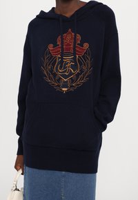 Sweat à capuche tricoté bleu marine avec un motif central brodé représentant une couronne et un crâne, poche kangourou, poignets et ourlet côtelés.