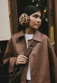 Brown faux leather jacket with a collar, button closure, and wide sleeves. Model wears leopard print earmuffs, a white shirt, and a crossbody strap.