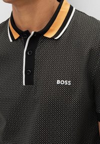 Polo shirt nero con motivo geometrico bianco, con collare a righe arancioni e nere e logo "BOSS" bianco.