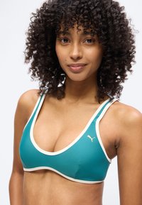 Noor da rohkete lokkidega, seljas sinakashall spordirinnahoidja valge äärega ja väikese Puma logoga rinnal, naeratab kerge naeratusega.