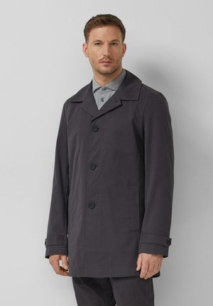 Man draagt een donkergrijze jas met knopen over een lichtgrijs overhemd met kraag, staand tegen een effen lichtgrijze achtergrond.