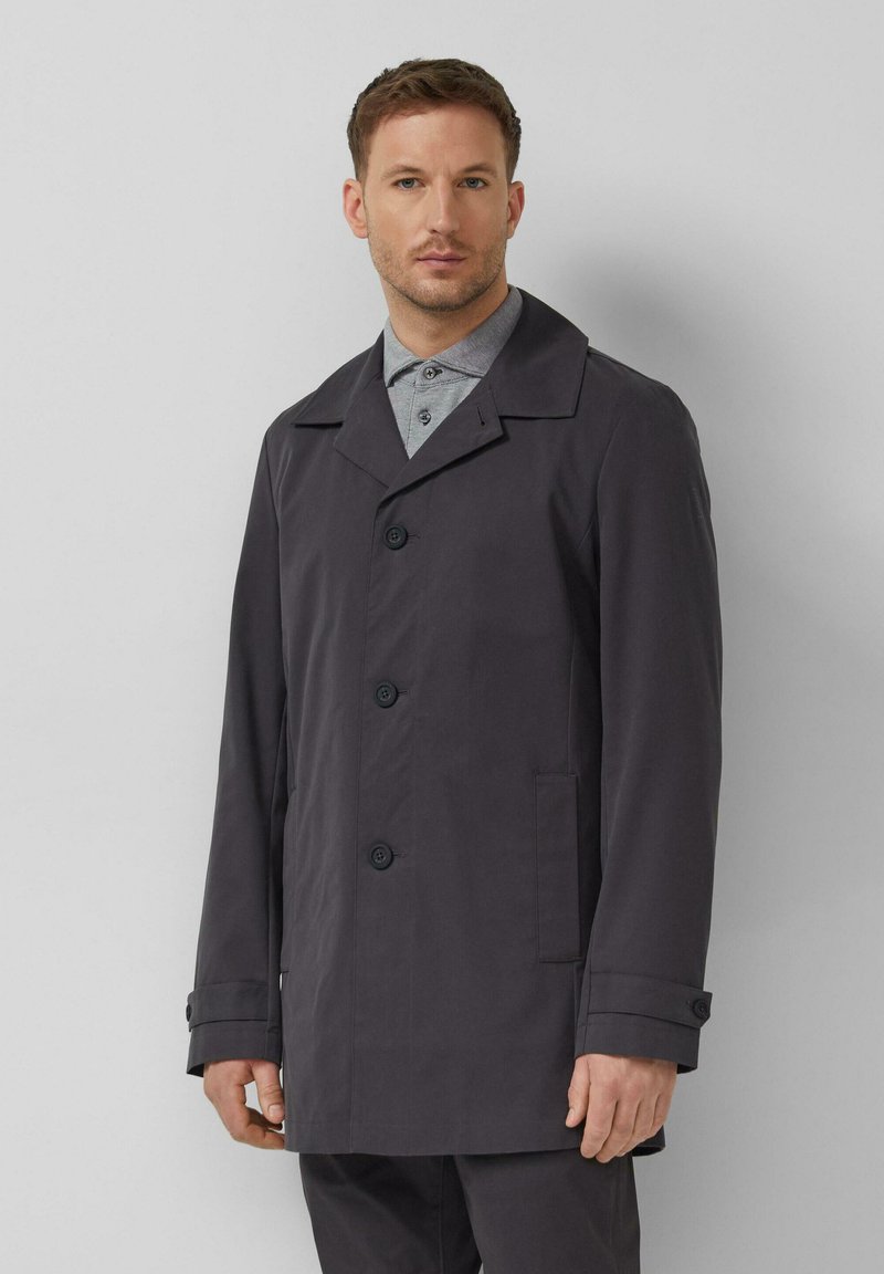 Mann trägt einen dunkelgrauen, geknöpften Mantel über einem hellgrauen Hemd mit Kragen und steht vor einem einfarbigen hellgrauen Hintergrund.