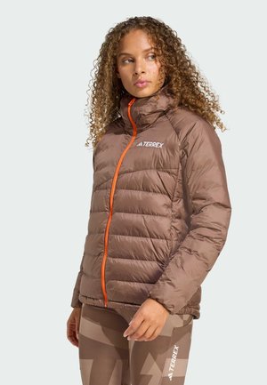 Kvinna med lockigt hår som bär en brun Terrex puffjacka med orange dragkedja och matchande mönstrade tajts, ser åt sidan.