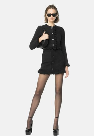 Completo in maglia nera con dettagli arricciati, composto da un cardigan corto e una gonna attillata con bottoni. Modella che indossa occhiali da sole e tiene una borsa.