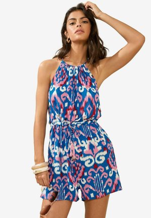 Jupe-culotte halter bleu coloré avec des motifs abstraits roses et blancs, taille cintrée et détails à franges. Les accessoires incluent des bracelets transparents.
