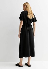 Vestido negro de longitud midi, hecho de tela texturizada, con mangas cortas y cintura ajustable. Llevado con sandalias planas negras.