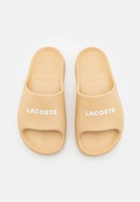 Sandalias tipo slip-on de Lacoste en beige, hechas de goma suave, con una apertura amplia, suela texturizada y un prominente logo blanco en la parte superior.