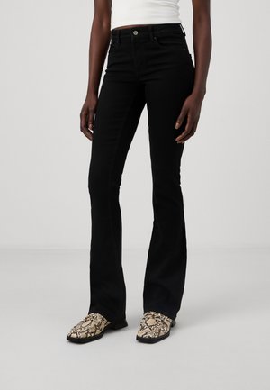 Femme portant un jean noir évasé taille haute et des bottines beige à motif peau de serpent, debout devant un fond clair uni.