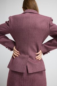 Vrouw met lang haar, gekleed in een paarse visgraat suit, die met haar handen op haar onderrug staat, gericht naar een effen lichtgrijze achtergrond.