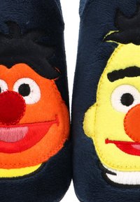 Zapatillas suaves de color azul oscuro con caras bordadas que tienen ojos grandes, narices rojas y cabello negro, una de color naranja y otra amarilla.