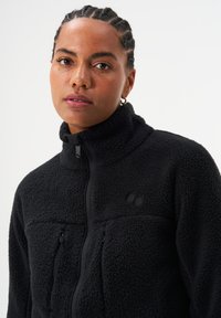 Schwarze Fleecejacke mit hohem Kragen, Reißverschluss und strukturierter Stoffoberfläche. Verfügt über zwei Fronttaschen und ein dezentes Logo auf der Brust.