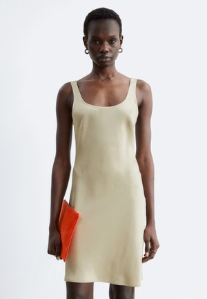 Frau trägt ein ärmelloses beiges Kleid und hält eine leuchtend orangefarbene Clutch vor einem schlichten, hellen Hintergrund.