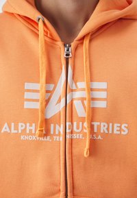 Narancssárga cipzáras kapucnis felső, fehér "ALPHA INDUSTRIES" logóval és húzózsinórral, amely puha tapintású és elöl cipzáras részlettel rendelkezik.
