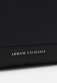 Sac en cuir noir avec une finition texturée. Présente le logo argenté "ARMANI EXCHANGE" affiché de manière proéminente à l'avant.