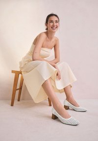 Witte open-toe schoenen met een geweven ontwerp en vierkante vorm, voorzien van een houten blokhak. Model draagt een lichtgekleurde strapless jurk en zit.