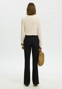 Femme aux cheveux mi-longs portant un haut crème à manches longues, un pantalon noir, tenant un sac à main tressé couleur tan, debout de dos.