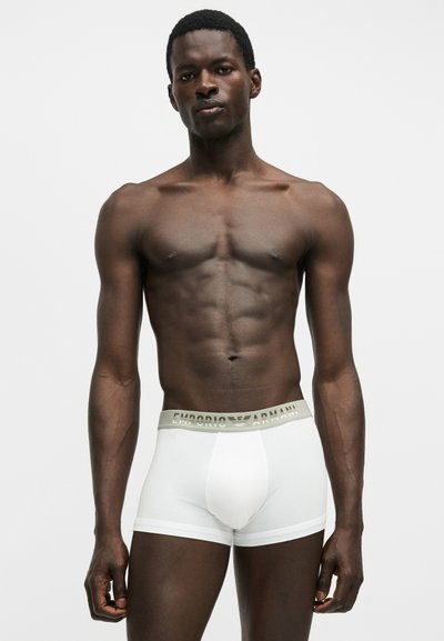 Μυώδης άνδρας στέκεται, φοράει λευκά boxer Emporio Armani με γκρι ζώνη, ουδέτερη έκφραση προσώπου σε απλό φόντο.