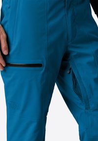 Detailní záběr osoby v modrých kalhotách North Face s kapsami na zip a síťovinovým ventilací na stehně.