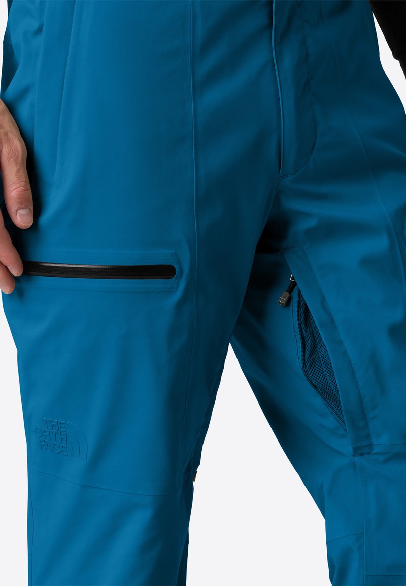 Primo piano di una persona che indossa pantaloni blu North Face con tasche con zip e ventilazione in rete sulla coscia.