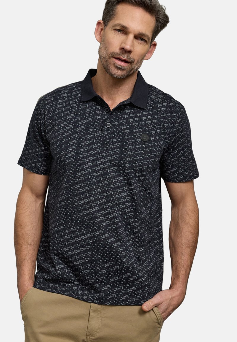 Homme portant un polo noir à motifs et un pantalon kaki, les mains dans les poches, debout devant un fond clair uni.