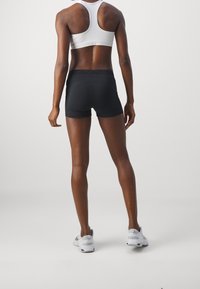 Pantalones cortos deportivos negros con diseño ajustado y textura suave; usados con un top deportivo blanco; muestra vista trasera, destacando un ajuste ceñido y detalles de la cinturilla.