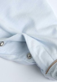 Lichtblauwe stof met een zachte textuur, voorzien van zilveren drukknoopjes en een gestikt bruin accent aan de zoom. Glad, lichtjes gekreukt uiterlijk.