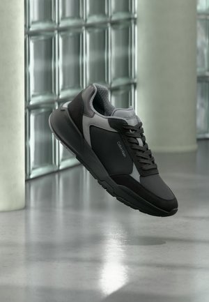 Μαύρο και γκρι αθλητικό παπούτσι Calvin Klein με κορδόνια, αιωρούμενο πάνω από ένα γυαλισμένο πάτωμα κοντά σε τοίχο από γυάλινα τούβλα και κολώνα από σκυρόδεμα.