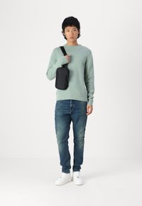 Light green sweater with ribbed cuffs, blue denim jeans, and white sneakers. Black crossbody bag rests on the shoulder. Minimalist design.