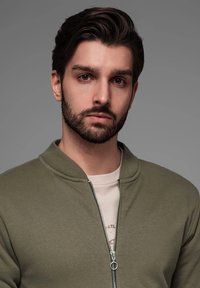 Giacca bomber verde con zip e colletto a costine, indossata sopra una maglietta chiara. Presenta un tessuto liscio e un design minimalista.