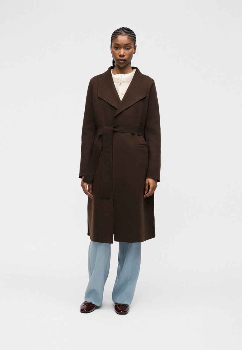 Γυναίκα όρθια φορώντας σκούρο καφέ παλτό με ζώνη, ανοιχτό μπλε παντελόνι, λευκό τοπ και σκούρα καφέ παπούτσια, μπροστά σε απλό ανοιχτό φόντο.