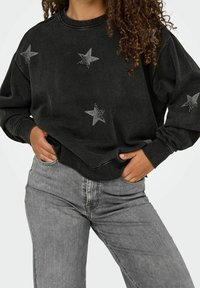 Sweat-shirt noir avec des embellissements en forme d'étoiles argentées, col rond, manches longues et coupe décontractée, associé à un jean gris clair.
