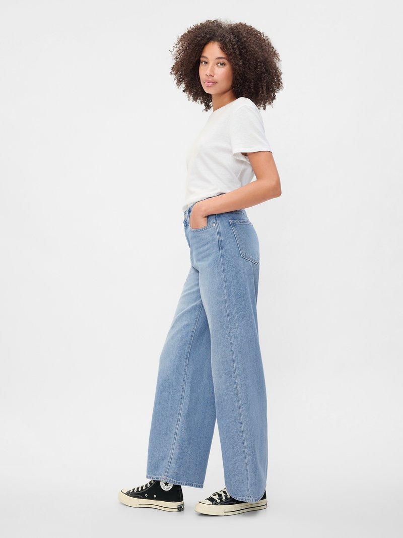 Frau mit lockigem Haar, die ein weißes T-Shirt, hellblaue weite Jeans und schwarze High-Top-Sneaker trägt, steht mit den Händen in den Taschen.