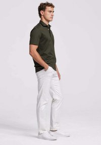 Donkergroene polo met korte mouwen en een kraag, gecombineerd met witte slim-fit broek en witte sneaker, staand tegen een effen achtergrond.