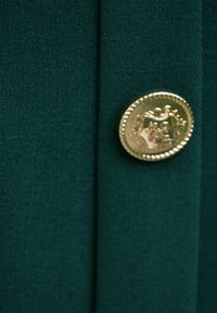 Zielona tkanina o gładkiej fakturze, z okrągłym złotym guzikiem wypukłym z dekoracyjnym wzorem, na tle czystym.
