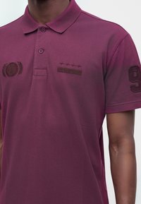 Homme portant un polo bordeaux avec logo Hollister brodé sur la poitrine et le numéro 9 sur la manche.