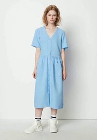 Jovem mulher a usar um vestido azul claro, com botão e de comprimento até ao joelho, mangas curtas, ténis brancos de cano alto e meias às riscas, de pé contra uma parede branca.