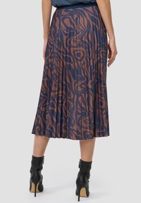 Žena v plisované midi sukni s abstraktním vzorem v námořnické modři a hnědé, s černými vysokými podpatky, viděná zezadu.