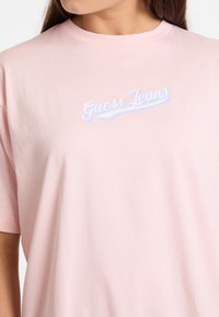 Lys pink bomulds t-shirt med korte ærmer og rund halsudskæring. Fronten viser teksten "Guess Jeans" i blå kursivskrivning.