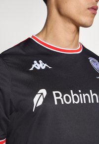 Camiseta deportiva negra con acentos rojos y textura horizontal, con los logotipos de Kappa en blanco y Robinhood en blanco, con cuello redondo.