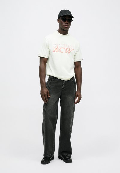 Camiseta verde claro con texto gráfico rojo, combinada con unos pantalones anchos de un gris oscuro y zapatos negros. Los accesorios incluyen gafas de sol negras y una gorra.