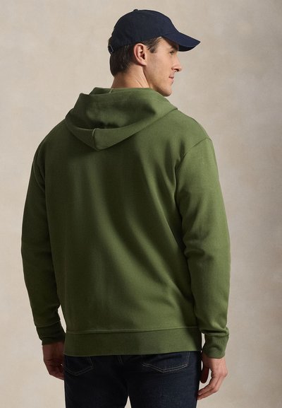 Mees, kes kannab oliivirohelise kapuutsiga pusa ja tumesinist nokamütsi, külje pealt, näidates pusa tagakülge ja osa tema näost.