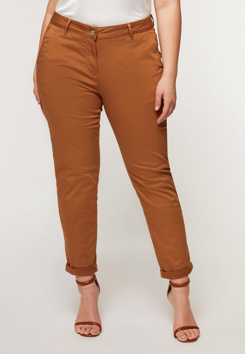 Femme portant un pantalon slim marron retroussé et des sandales à talons hauts marron à lanières, sur un fond neutre.