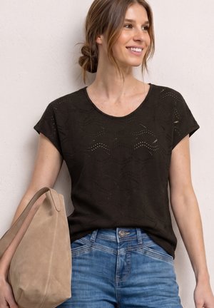 Smilende kvinde med brunt hår i en sort mønstret kortærmet top og blå jeans holder en beige skuldertaske op ad en bleg væg.