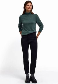 Dunkelgrüner Rollkragenpullover aus weichem Strickstoff, kombiniert mit schwarzen Slim-Fit-Hosen. Schwarze Loafers vervollständigen das Outfit.