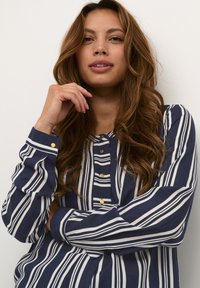 Blusa a rayas azul marino y blanca con frente de botones, cuello redondo y mangas largas con puños abotonados. Textura de tela suave.