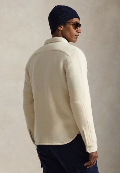 Muž v krémovom vlnenom saku, tmavomodrej pletenej čiapke, slnečných okuliaroch a tmavých nohaviciach stojaci z profilu na jednoliatom béžovom pozadí.